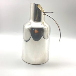 Könnte beinhalten: Eine silberne Kanne mit einem einzigartigen, gebogenen Griff. Die Kanne hat eine glatte, polierte Oberfläche und ein modernes Design.