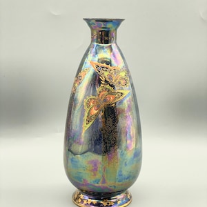 Op de afbeelding: Een peervormige iriserende vaas met een uitlopende hals en een goudkleurige voet. De vaas is versierd met goudkleurige vlinders. De vaas heeft een donkerblauw, paars en goud kleurenschema.
