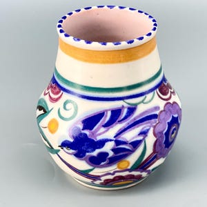 May include: A small, cream-colored ceramic vase with a rounded body and a flared rim. The vase features a hand-painted design of a blue bird, purple flowers, and green leaves. The rim is pink with blue dots, and there is a yellow band.