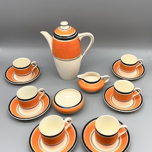May include: A vintage orange and white ceramic tea set. The set includes a tall coffee pot, several cups and saucers, a creamer, and a sugar bowl. Each piece features orange, black, and gray stripes. The set is arranged on a gray surface.