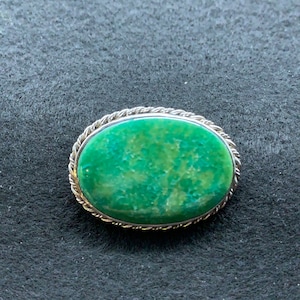 May include: A silver brooch with a green oval stone set in a rope-like border.