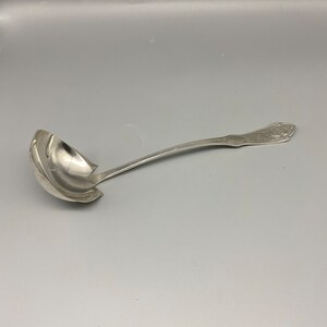 Könnte beinhalten: Eine silberne Schöpfkelle mit einem verzierten Griff. Die Kelle hat eine tiefe Schale und einen langen, gebogenen Griff.