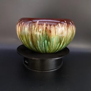 May include: A decorative ceramic bowl with a rounded shape. The bowl features a glossy finish with a color gradient, transitioning from a dark brown rim to green and gold tones. The bowl is displayed on a black stand.