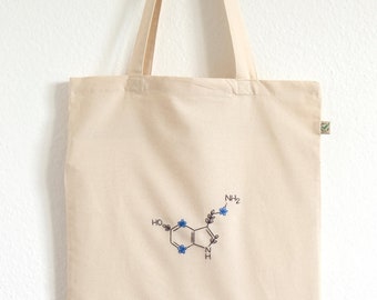 May include: A beige canvas tote bag with a chemical structure embroidered on the front. The structure is of serotonin, a neurotransmitter, and is decorated with blue flowers. The bag has a green tag on the side.