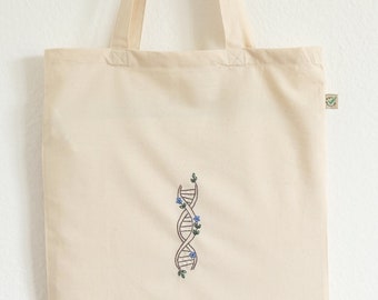 May include: A natural canvas tote bag with a simple embroidered design of a DNA strand with blue flowers. The bag has a green eco-friendly label.