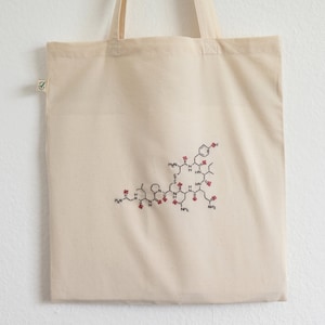 May include: Beige canvas tote bag with a black and red embroidered molecular structure design. The bag has two handles and a small green tag. The design is centered on the front of the bag.