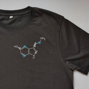 Może przedstawiać: Ciemnoszary t-shirt z krótkim rękawem z wyhaftowaną białą strukturą chemiczną, ozdobioną małymi jasnoniebieskimi kwiatowymi detalami. Wzór chemiczny zawiera tekst HO, NH2 oraz N, z delikatnymi motywami liści wkomponowanymi w całość projektu.