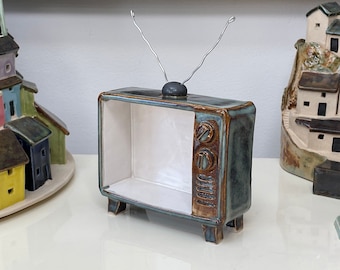 Può includere: Una scultura di televisione retrò in ceramica con una glassa verde acqua e marrone. La TV ha un interno bianco, due manopole e un paio di antenne a filo. Case decorative in ceramica sullo sfondo.
