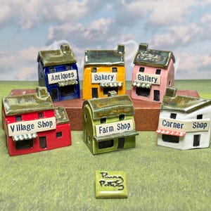 May include: A selection of miniature ceramic shop buildings in various colours, including red, blue, yellow, pink, green, and white. Each building has a sign indicating its type, such as "Antiques" or "Bakery". A small tile with the text "Penny Pottery" is also present.