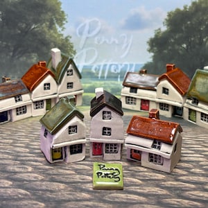 Op de afbeelding: Een verzameling miniatuur keramische huisjes met verschillende dak kleuren, waaronder rood, groen en bruin. De huisjes hebben witte muren, kleine ramen en schoorstenen. Er is ook een kleine tegel met de tekst "Penny Pottery".