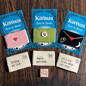 Könnte beinhalten: Drei keramische, umschlagförmige Ornamente mit dem Text "Kerenza Sent in Stone" auf blauen Karten. Die Umschläge sind rosa, grün und schwarz, mit Herz-, Pfotenabdruck- und Raketenmotiven. Darunter befinden sich drei kleinere Ornamente mit dem Text "Sending you love", "You're purr-fect" und "Love you to the moon and back".