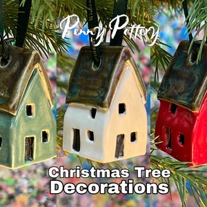 May include: Three ceramic house ornaments, one green, one white, and one red, with brown roofs and black window details. The ornaments are hanging from a Christmas tree and are labeled "Christmas Tree Decorations".