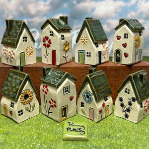 May include: A collection of ten miniature ceramic houses with green roofs and colourful floral details. Each house has a unique design and features a small door and windows. The houses are arranged on a wooden surface with a green grassy background.