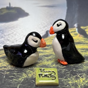 May include: Two ceramic puffin figurines, one sitting and one standing, with black and white bodies, orange beaks and feet. A small green tile with the text "Penny Potter" is in front of them. The background shows a coastal scene with a small island.