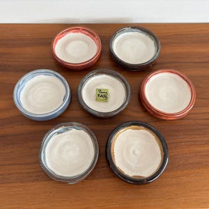 Könnte beinhalten: Sieben kleine, runde Keramikschalen mit weißem Innenraum und unterschiedlich farbigen Rändern in Blau, Rot, Braun und Schwarz. Eine Schale in der Mitte enthält ein kleines grünes quadratisches Plättchen mit der Aufschrift Penny Pottery.