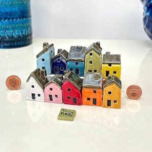 May include: A collection of ten miniature ceramic houses in various colours including white, pink, red, orange, yellow, light blue, dark blue, teal, and green. Each house features a dark grey roof, a chimney, and small square windows and doors.