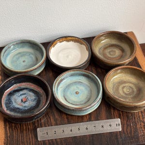 May include: Six small ceramic bowls in shades of blue, brown, and white sit on a wooden surface. A ruler with centimetre markings is placed near the bowls. The bowls have a rustic, handmade appearance, suggesting artisanal craftsmanship.