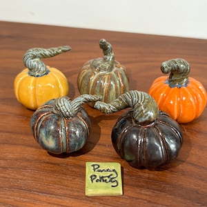 May include: Five small ceramic pumpkins with textured stems in yellow, green, orange, blue-grey, and dark brown glazes. A small square tile in front features the handwritten text "Penny Pottery" in black ink on a light green background.