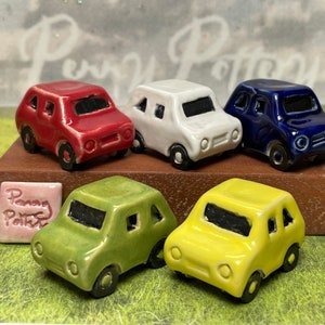 Puede incluir: Cinco coches en miniatura de cerámica en diferentes colores: rojo, blanco, azul, verde y amarillo. Los coches están dispuestos en fila sobre una superficie marrón. El texto "Penang Pottery" es visible en un azulejo rosa.