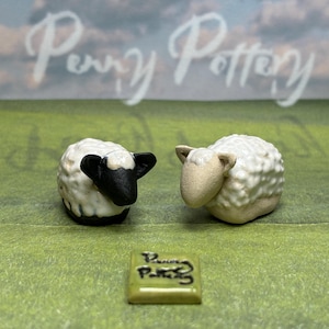 May include: Two ceramic sheep figurines with textured white wool and black and tan faces. A small, square tile with the text "Penny Pottery" is in front of the sheep. The background is a green field with a cloudy sky.