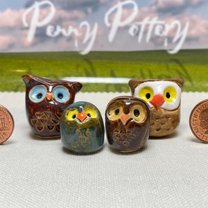 Może przedstawiać: Cztery małe ceramiczne figurki sów, każda z różnymi kolorami oczu i wzorami. Sowy są brązowe, zielone i białe z pomarańczowymi dziobami. Sowy siedzą na białym tle obok amerykańskiego centa i brytyjskiego pensa.