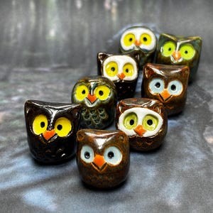 May include: A collection of eight ceramic owl figurines in various shades of brown, green, and white. The owls have yellow eyes and orange beaks.