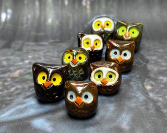 Puede incluir: Una colección de ocho figuritas de búho de cerámica en varios tonos de marrón, verde y blanco. Los búhos tienen ojos amarillos y picos naranjas.