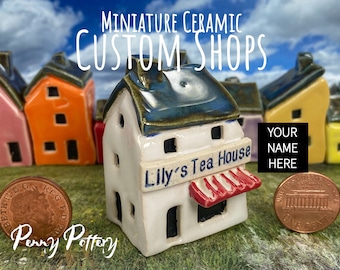 May include: A miniature ceramic shop building with a white exterior, blue roof, and red awning. The shop is labelled "Lily's Tea House". The building is surrounded by other miniature ceramic shops in various colours. A penny is placed in front of the building for scale.