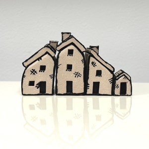 May include: A set of four miniature houses made of paper, painted in a light beige color with black details. The houses are arranged in a row, with the largest house in the center and the smallest house on the right.