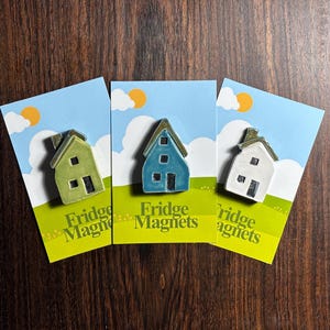 May include: Three fridge magnets featuring miniature ceramic houses in green, blue, and white. Each house has a contrasting roof and black windows and door. The magnets are on cards with a sky and grass design, and the text "Fridge Magnets".