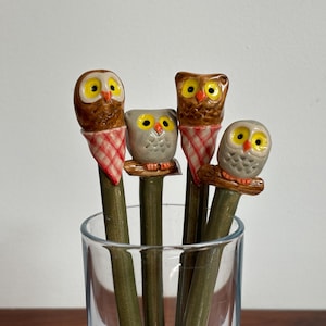 May include: Four decorative pencils with owl toppers in a clear glass. The owl toppers are brown, gray, and white with yellow eyes. Each owl is perched on a green pencil. The pencils are arranged in a clear glass.