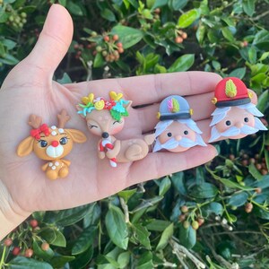 May include: Four handmade polymer clay Christmas ornaments. A red and white reindeer, a brown deer with a green and gold wreath, a white bearded man with a red and white hat, and a white bearded man with a red and gold hat.
