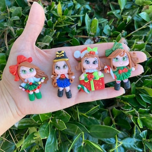 May include: Four handmade Christmas-themed polymer clay ornaments featuring girls wearing festive attire. The ornaments are decorated with details like bows, candy canes, and presents.