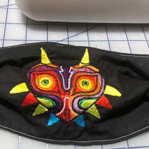 May include: Black fabric face mask with a colorful embroidered design of the Majora's Mask from the Legend of Zelda video game series.