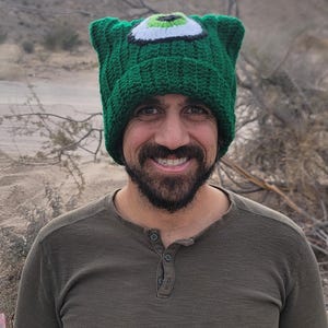May include: A person wearing a green knitted hat with a large green eye on the front. The hat has a white outline around the eye and a black pupil.