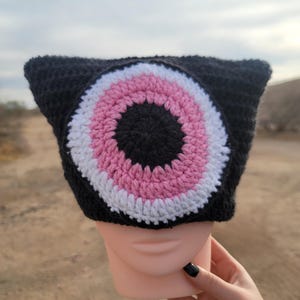 May include: Black crochet beanie with a large pink, white, and black eye design.