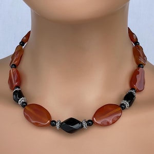 May include: A necklace featuring large, polished, oval-shaped orange-brown stones interspersed with smaller black beads and silver-toned accents. The necklace has a striking design with a mix of colors and textures, suitable for adding a bold statement to any outfit.