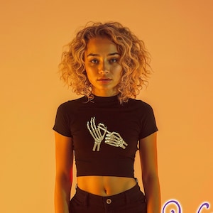 May include: A black crop top with a skeleton hand graphic. The top has a mock neck and short sleeves. The model is wearing black shorts.