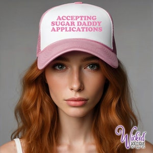 May include: A pink and white trucker hat with the text "ACCEPTING SUGAR DADDY APPLICATIONS" in pink letters. The hat is worn by a person with long red hair.