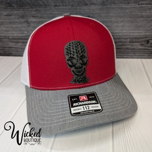 May include: Red, white, and gray trucker hat with a black skull graphic wearing sunglasses. The hat has the text "EST. 1970 RICHARDSON. ORIGINAL 112 TRUCKER ADJUSTABLE" on the back.