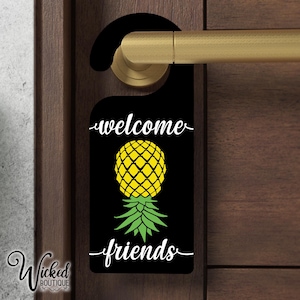 May include: Black door hanger with the text "welcome" and "friends" in white cursive font. A yellow pineapple with green leaves is in the center of the hanger.