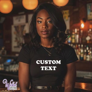 Puede incluir: Camiseta negra con las palabras "CUSTOM TEXT" en blanco. La modelo lleva un collar dorado y un cinturón marrón. El fondo es un ambiente de bar borroso con iluminación cálida.