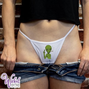 Pode incluir: Tanga branco com um desenho de um alienígena verde fumando um cigarro e mostrando o dedo do meio. O alienígena está vestindo uma camisa verde. O texto "Wicked Boutique" está em roxo.
