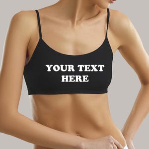 May include: Black sports bra with thin straps and the text "YOUR TEXT HERE" in white. The bra is worn by a person. The background is a solid light gray.