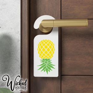 May include: White door hanger with a pineapple design. The pineapple is yellow with green leaves. The hanger is attached to a door with a gold handle.
