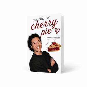 May include: A white greeting card featuring a smiling person with dark curly hair wearing a black long-sleeve shirt. The text reads YOU'RE MY cherry pie - GARRETT GRAHAM. A hand-drawn illustration of a slice of cherry pie with a heart is included.