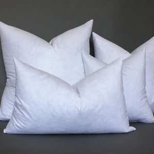 May include: Four white down pillows of different sizes stacked on top of each other against a gray background.