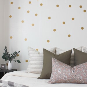 May include: A white bed with a white duvet and a gray and white throw blanket. The bed has a variety of pillows in different colors and patterns, including a striped pillow, a solid green pillow, and a floral pillow. The wall behind the bed is white with gold polka dots.