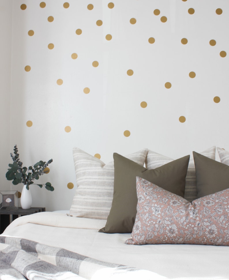 May include: A white bed with a white duvet and a gray and white throw blanket. The bed has a variety of pillows in different colors and patterns, including a striped pillow, a solid green pillow, and a floral pillow. The wall behind the bed is white with gold polka dots.