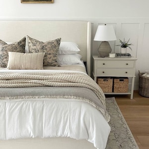 May include: A white bed with a beige headboard and a white duvet cover. The bed has a beige knitted throw blanket and a gray throw blanket with fringe. There are two pillows with a floral pattern and a beige pillow with stripes on top of the bed. A white lamp with a beige shade is on a nightstand next to the bed. The nightstand has two drawers and two wicker baskets. A small potted plant is on the nightstand.